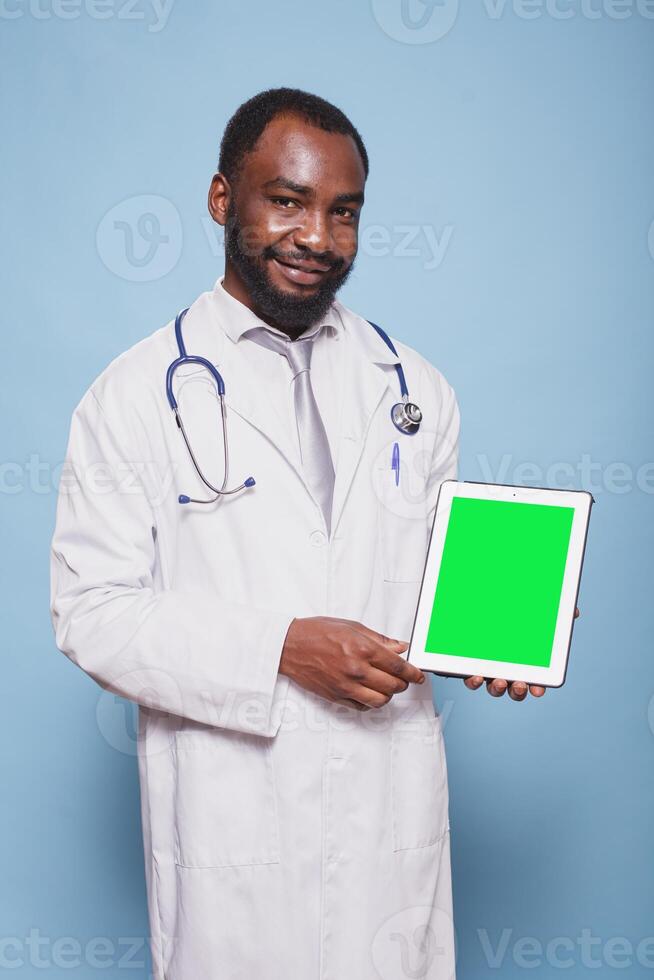 Portrait of black man wearing a stethoscope and lab coat is holding a tablet with green screen. African american doctor grasping device displaying blank chromakey mockup template. photo