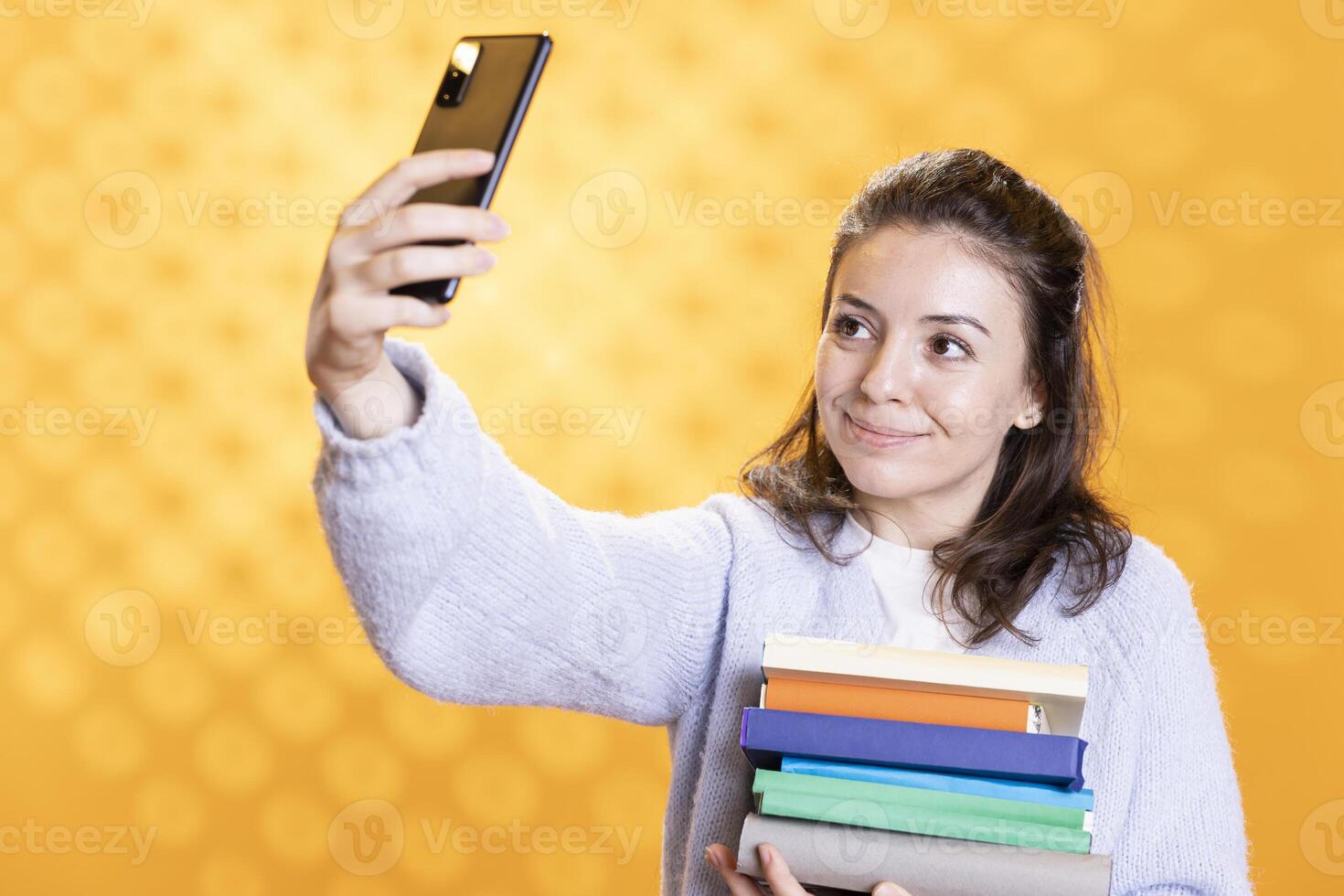 Smiling woman doing selfie with cellphone holding stack of books, isolated over studio background. Happy student with pile of textbooks in arms taking picture with mobile phone photo