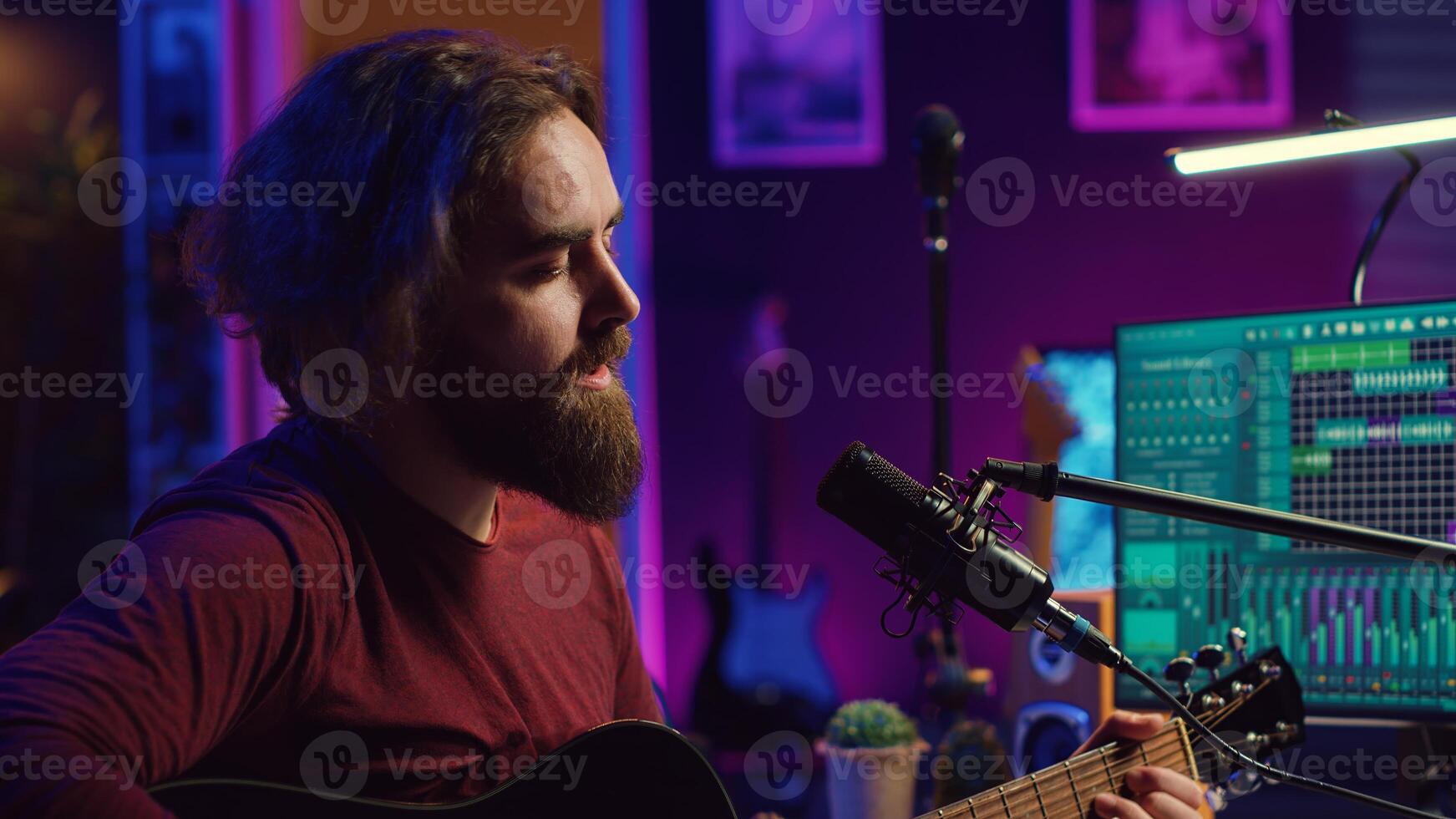 Songwriter producing tunes on acoustic guitar in his personal studio, using a microphone to sing the chords. Audio engineer works with soundboard and equalizer to compose new music. Camera A. photo