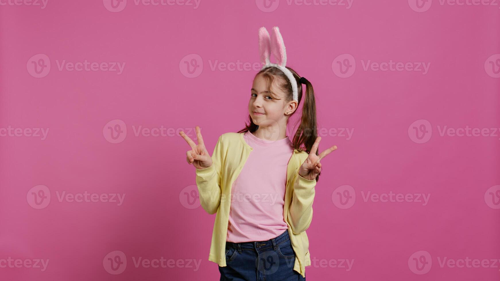 Young cheerful kid with pigtails showing peace sign in studio, feeling excited and confident about easter sunday celebration. Smiling youngster does signs against background. Camera B. photo