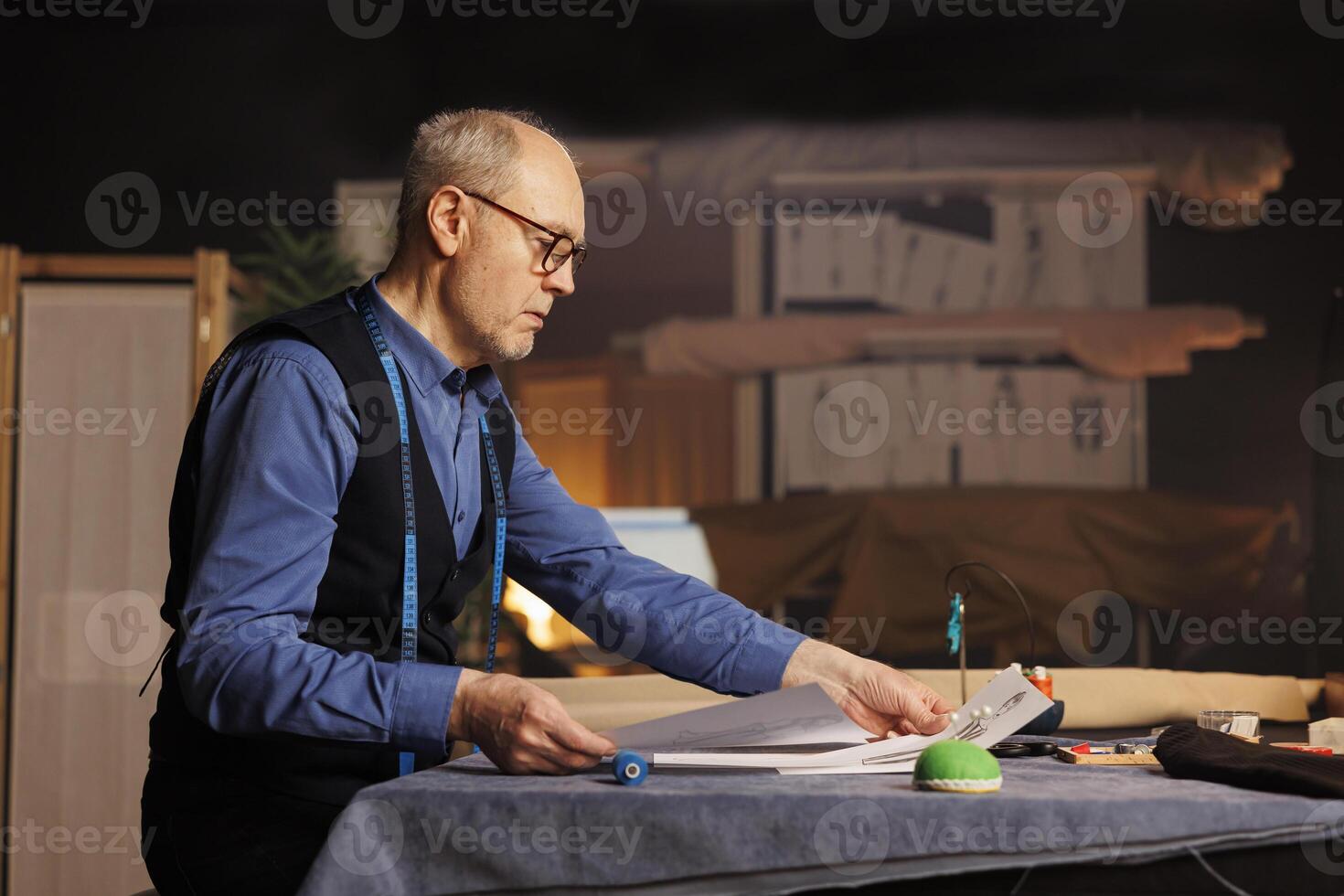 Couturer doing needlework in atelier shop workspace, checking bespoken clothing drafts. Suitmaker looking over sketch drawings, preparing to manufacture sartorial garments for client photo