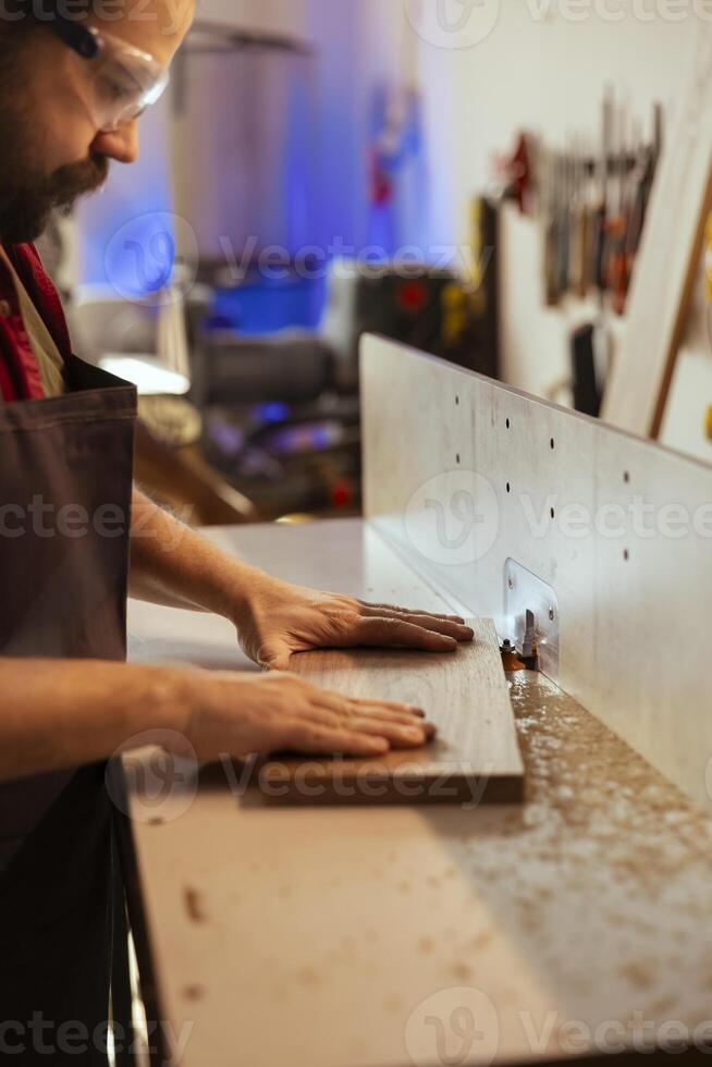 Man wearing protection equipment while inserting plank in spindle moulder to prevent workplace accidents. Carpenter equipped with safety glasses while cutting lumber with wood shaper to avoid injury photo