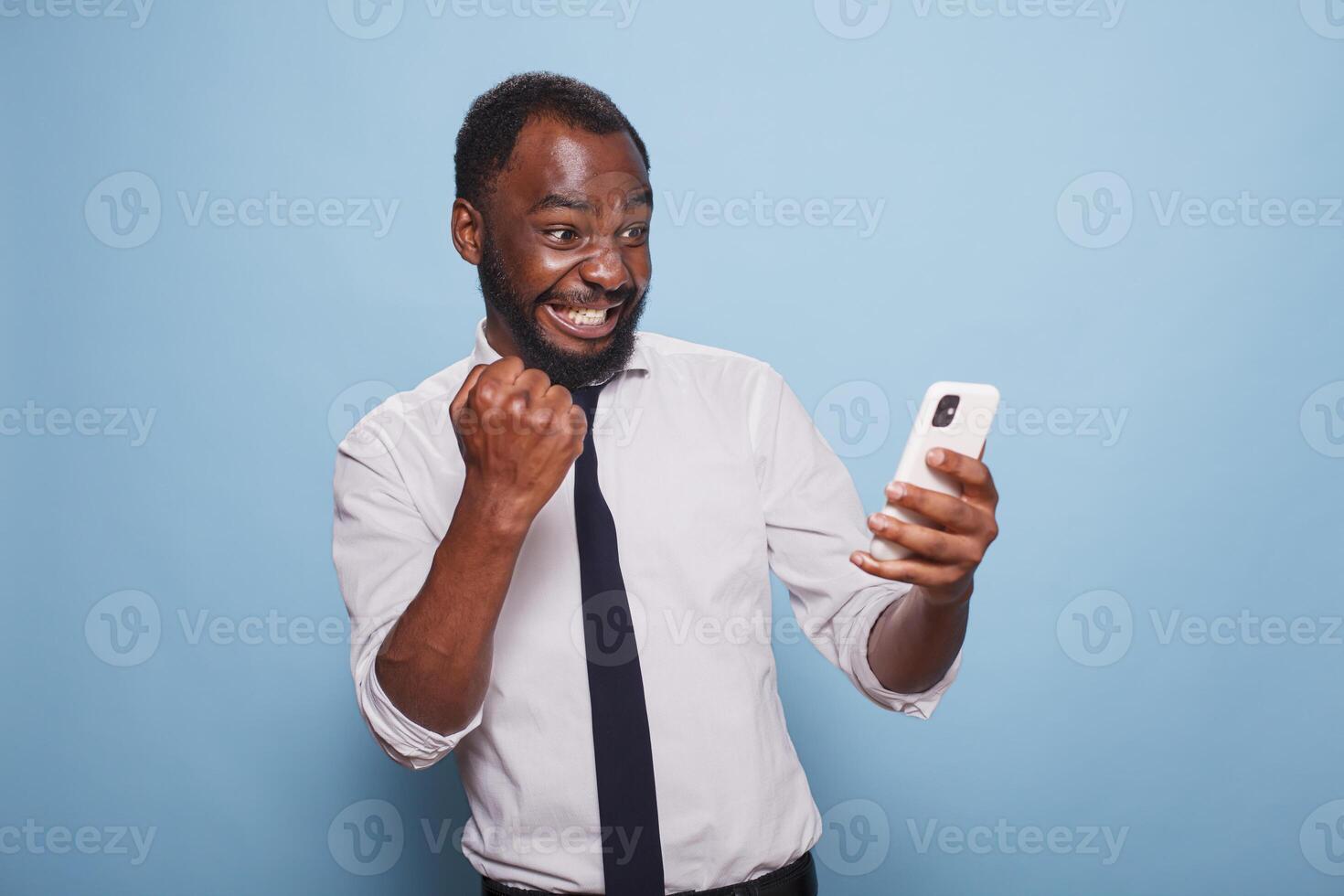 Black man in white shirt holding cellphone winning online mobile game looking tensed at screen. African american gamer testing mobile app on smart phone for the first time feeling enthusiastic. photo