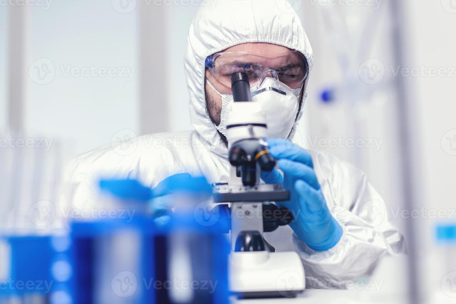 Microbiologist researcher wearing ppe and protection glasses in medicine lab looking through microscope. Scientist in protective suit sitting at workplace using modern medical technology during global epidemic. photo