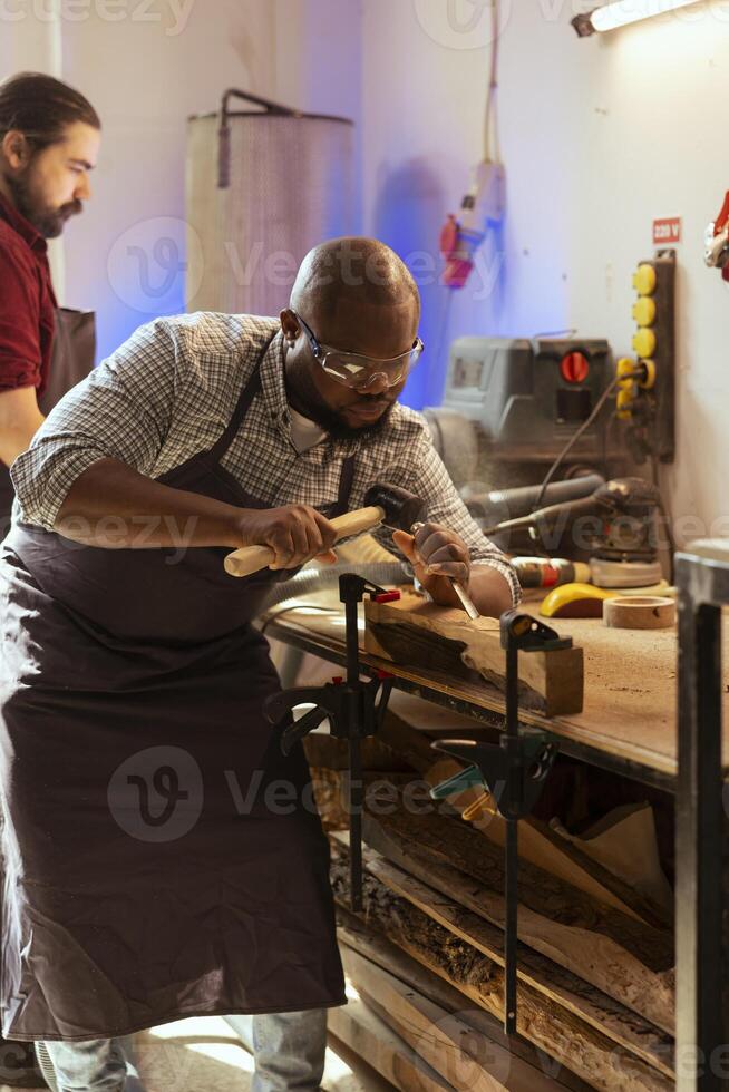 African american manufacturer carving intricate designs into wood using chisel and hammer in carpentry shop. BIPOC artisan in woodworking workshop shaping wooden pieces with tools, close up shot photo