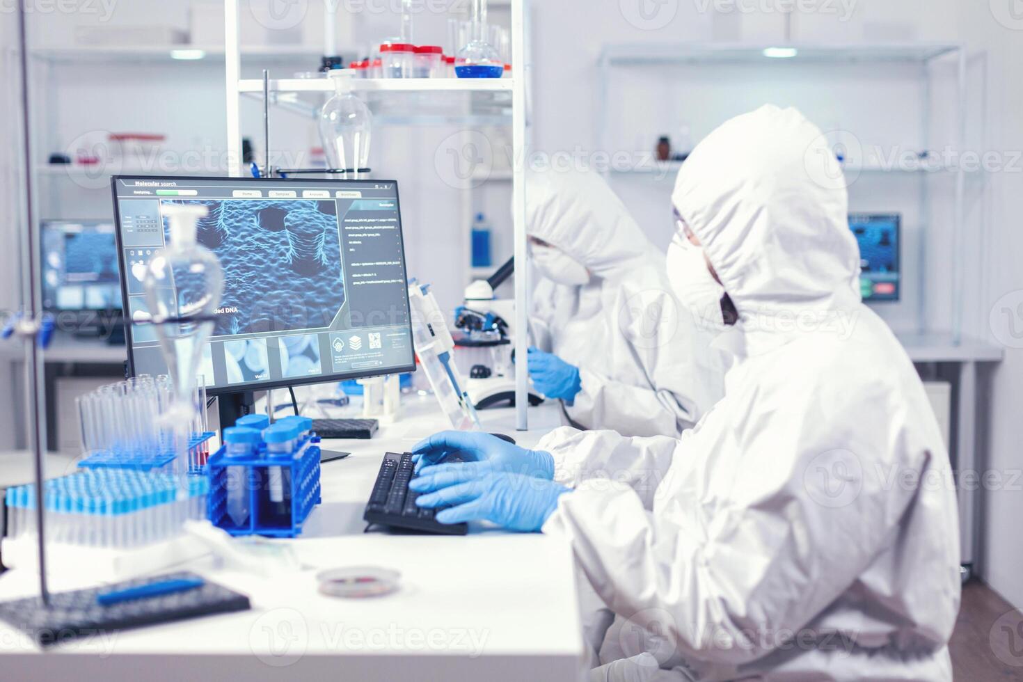 Lab engineer typing on pc conducting experiment in laboratory dressed in ppe suit. Medical engineer using computer during global pandemic with coronavirus dressed in coverall. photo