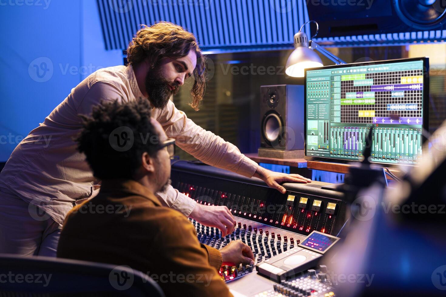 Skilled artist working with audio technician to edit recorded songs in control room, pressing sliders and levers on panel board. Team of singer and sound designer creating records in studio. photo
