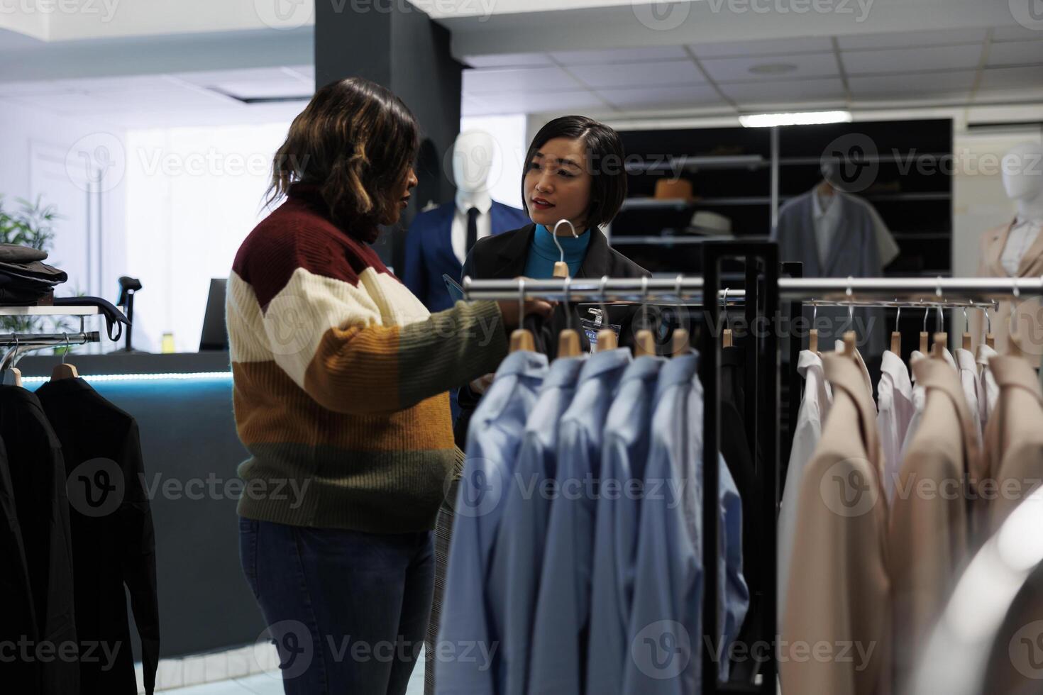 Clothing store woman asian assistant talking and consulting customer. Customer exploring rack with apparel, choosing formal outfit and getting help from boutique employee photo