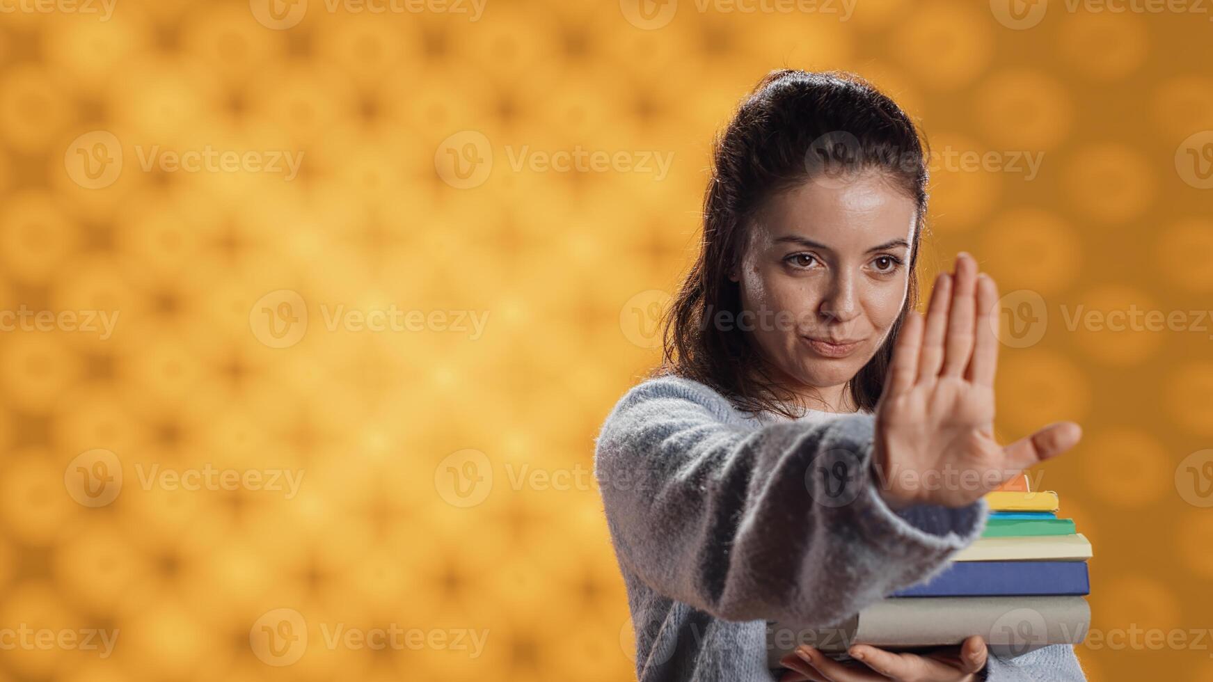Portrait of stern woman holding stack of books doing stop sign gesturing, studio background. Student with pile of textbooks in arms used for academic learning doing halt hand gesture, camera A photo
