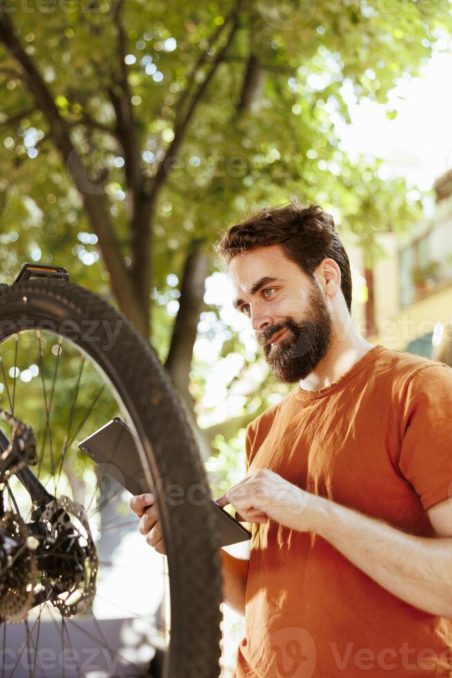Enthusiastic healthy male cyclist checking tire as yearly bike rubber maintenance routine with digital tablet. Caucausian man using smart smart device to look for solutions of damaged bicycle tire. photo