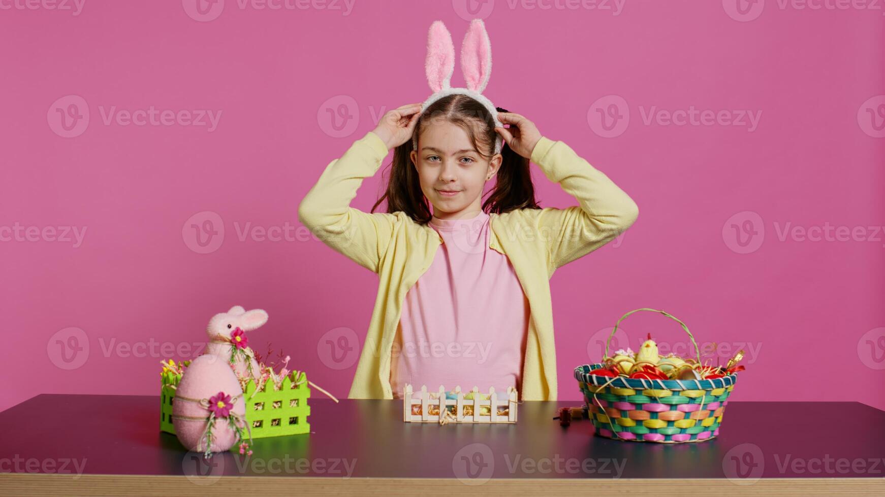 energético joven niña con adorable conejito orejas ondulación en estudio, diciendo Hola y saludo alguien mientras ella crea Pascua de Resurrección decoraciones alegre niñito posando en contra rosado fondo. cámara b. foto