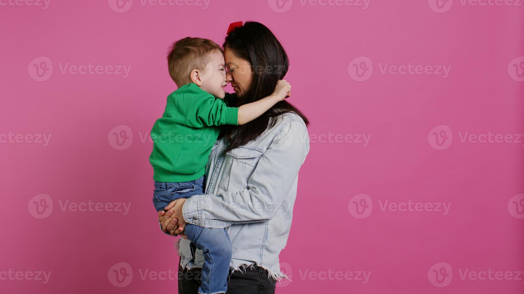 Adorable small boy hugging his mother and playing around, having fun against pink background. Young toddler posing with his mom holding him, laughing and being cheery. Camera B. photo