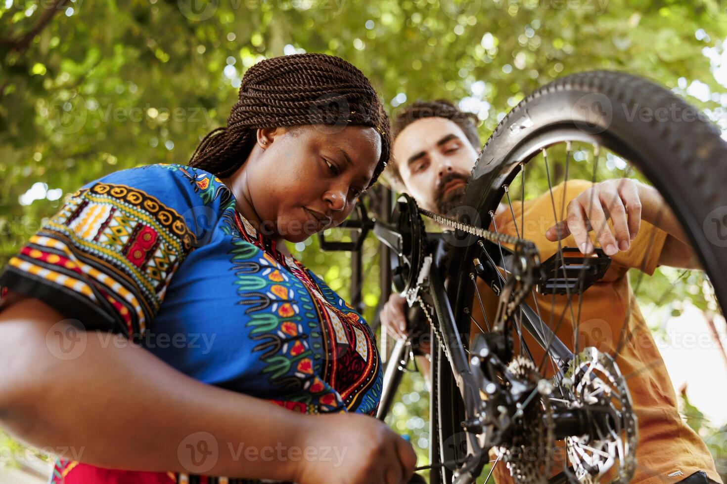 Sporty and eager black woman in yard repairing bike derailleur for summer recreational cycling. Healthy committed african american female cyclist assisted by caucasian man in repairing of bicycle components. photo