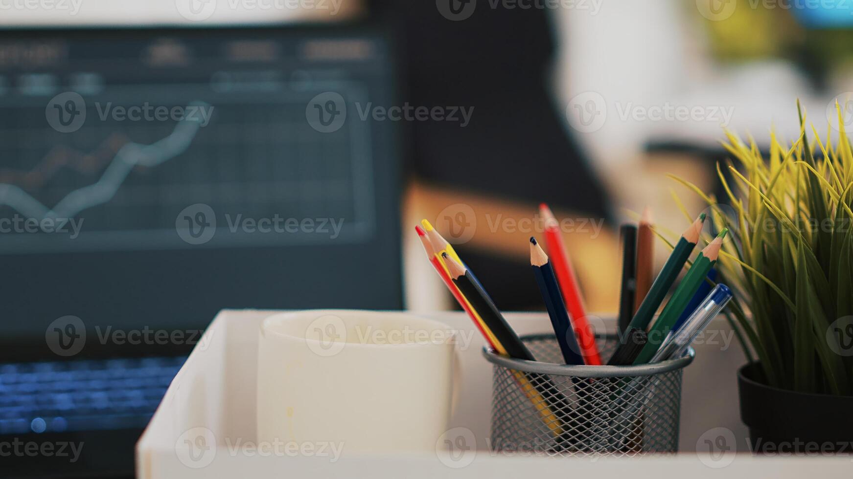 Focus on desk belongings in modern office with notebook in blurry background showing business diagram and figures, close up. Pencil holder, mini house plant, empty coffee mug and graphs on laptop photo
