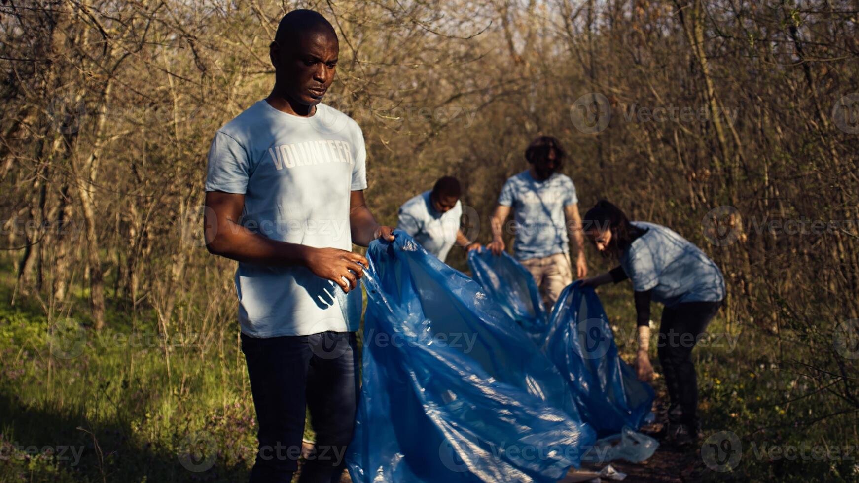Diverse volunteers collecting garbage and junk from the forest area, fighting environmental pollution and conservation. Group of activists cleaning up the woods, recycling plastic waste. Camera B. photo