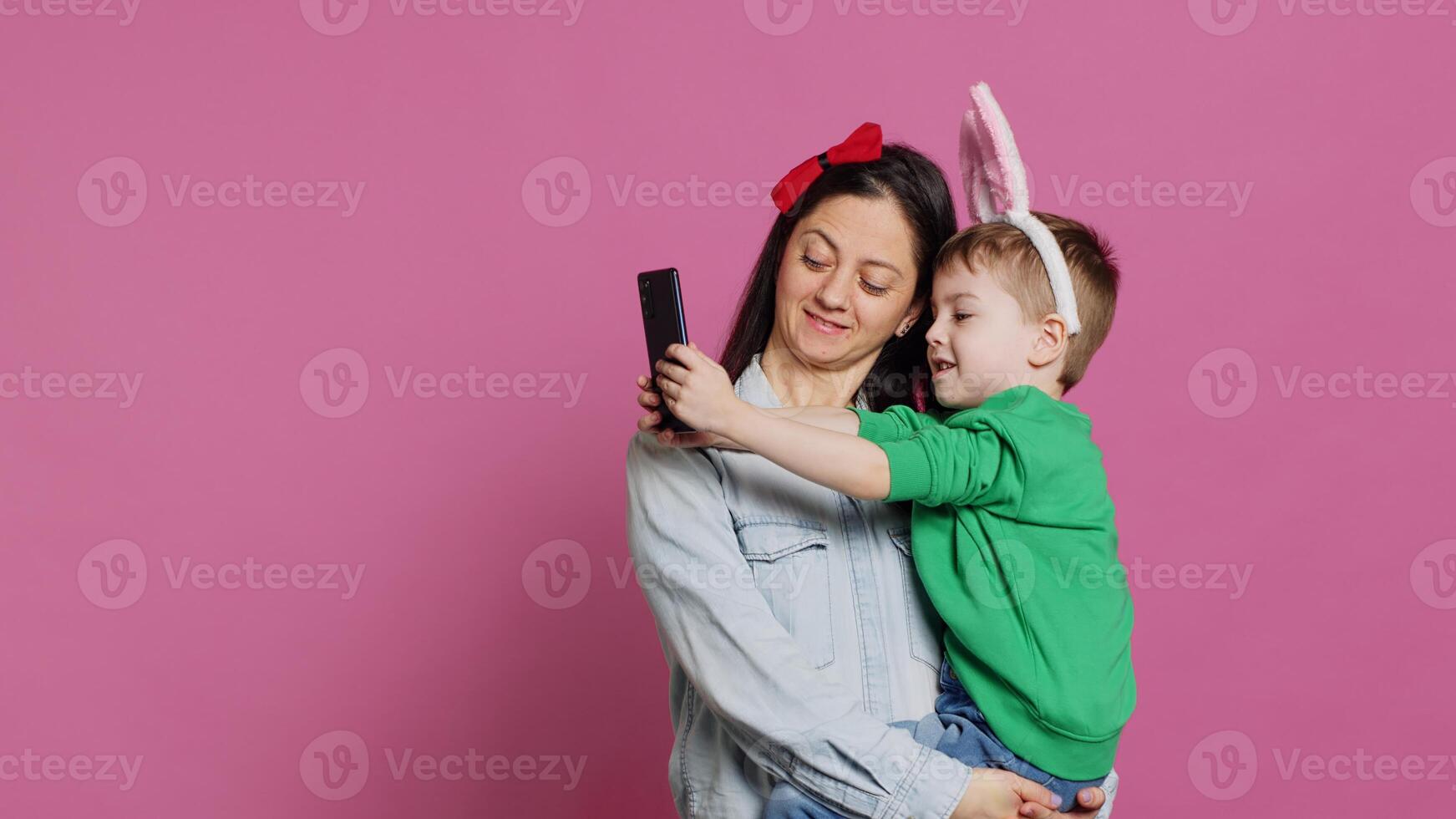 Lovely small child taking pictures with his mother on smartphone, trying to capture fun and cute moments against pink background. Little boy being playful and fooling around with phone. Camera A. photo