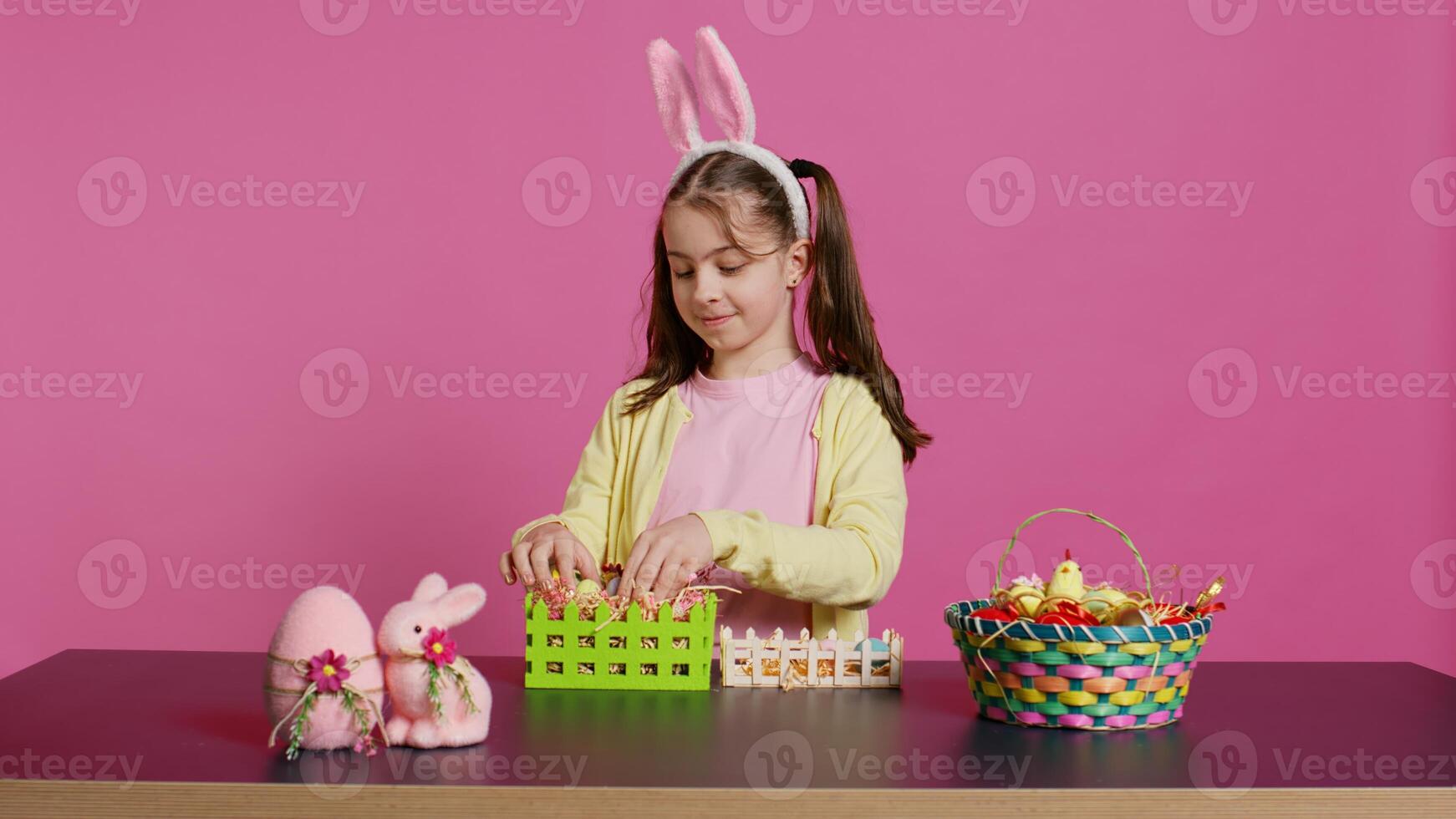 alegre pequeño niña demostración su hecho a mano festivo cesta en cámara, creando un vistoso hecho a mano arreglo en hora para Pascua de Resurrección día festivo. pequeño niñito con conejito orejas decorando huevos. cámara b. foto