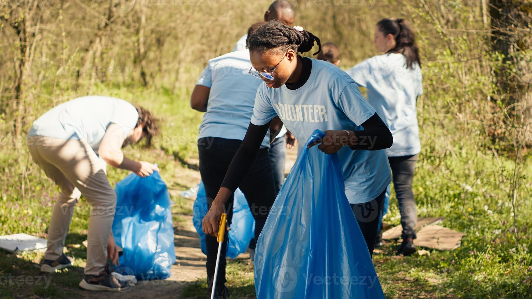 africano americano niña cosecha arriba basura con un largo garra y basura bolsas, limpieza bosque habitat y luchando ilegal dumping con un equipo de voluntarios activista coleccionar basura. cámara una. foto