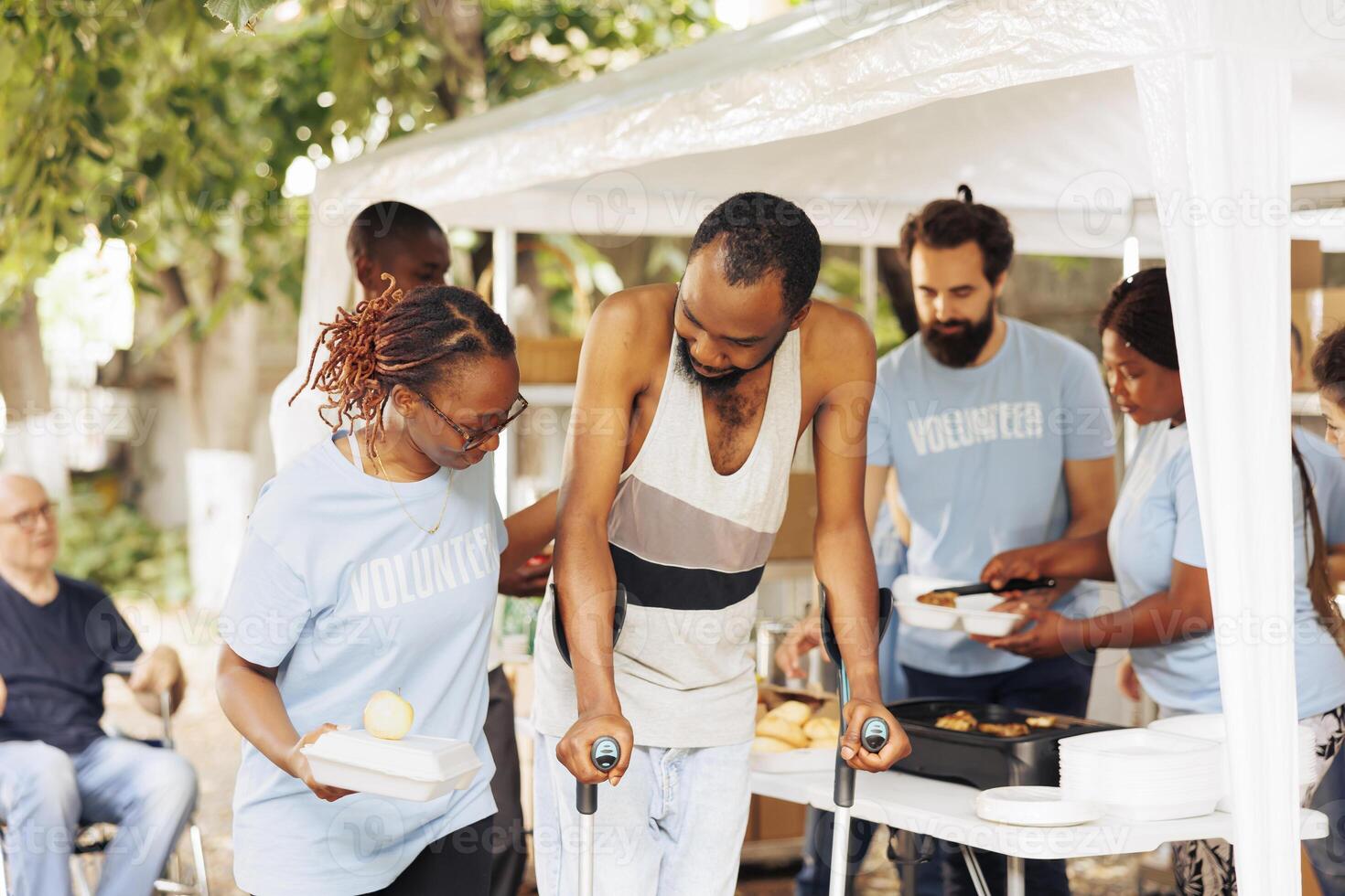 Multiethnic charity group handing out free food and nourishments to less fortunate and disabled. Portrait of black woman with blue t-shirt assisting the poor, needy african american man on crutches. photo
