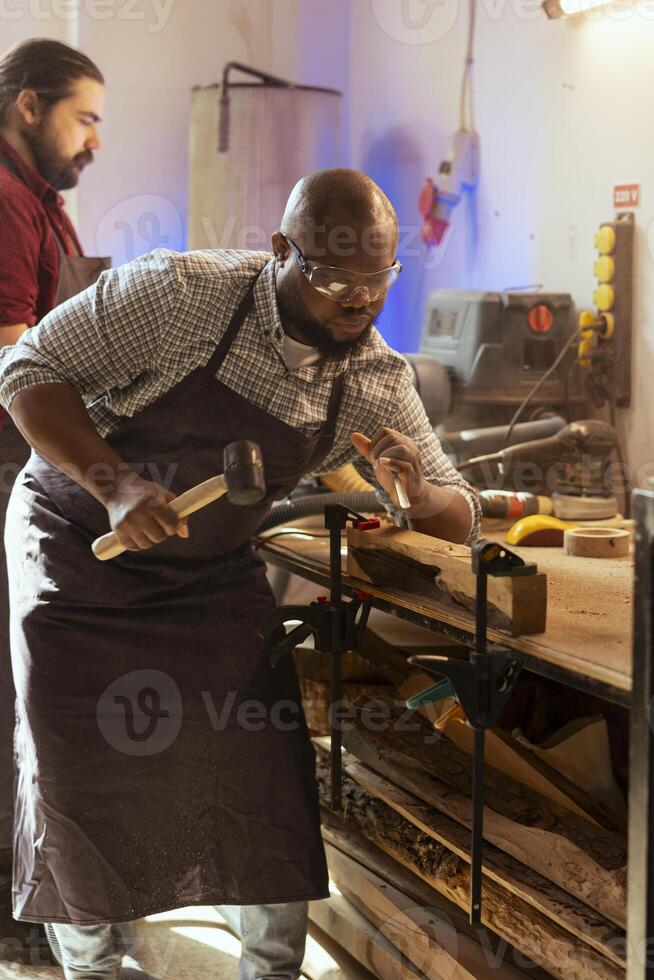 artesano tallado diseños dentro madera utilizando cincel y martillo siguiente a compañero de trabajo artesano formación de madera piezas utilizando protector equipo a evitar lugar de trabajo accidentes, ayudó por aprendiz foto