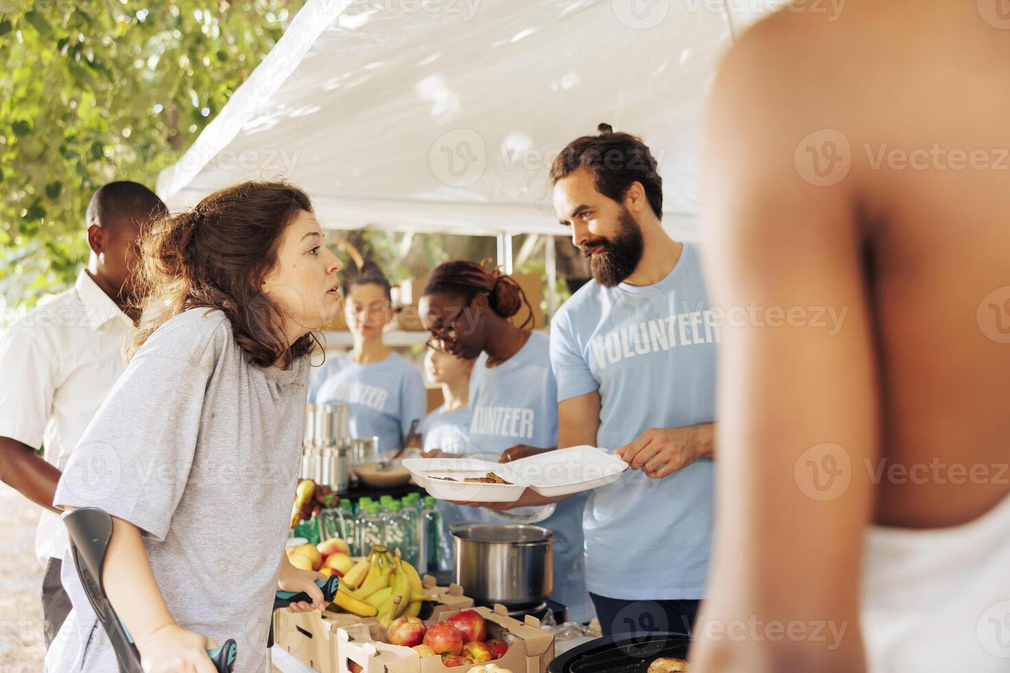 Image shows caucasian woman on crutches receives assistance from multicultural volunteers for hunger relief. Free food and humanitarian aid provided to needy and crippled by non-profit organization. photo