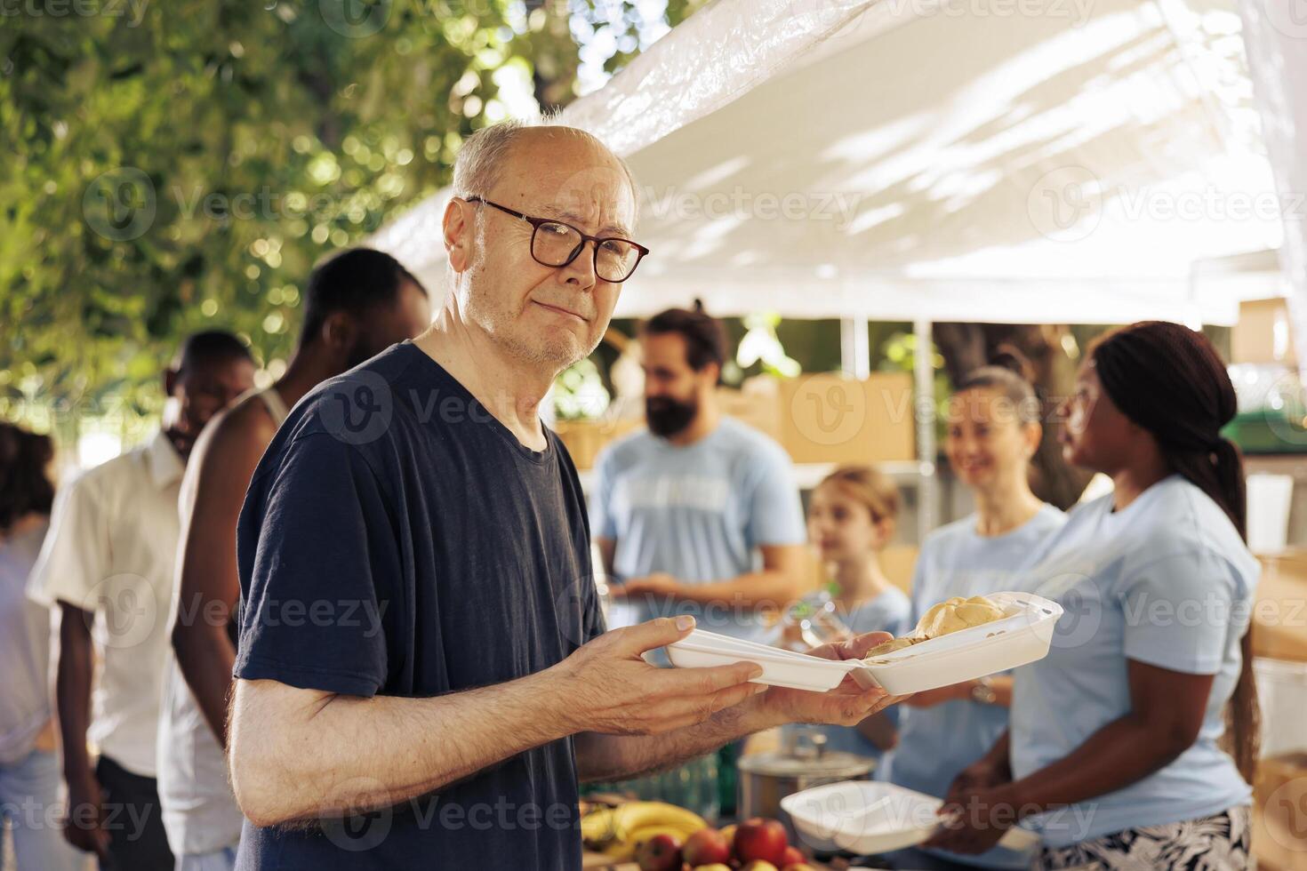Image showcasing old caucasian homeless man holding his meal from the charitable organization. Group of volunteers at an outdoor food bank helping and feeding the hungry poor and less privileged. photo