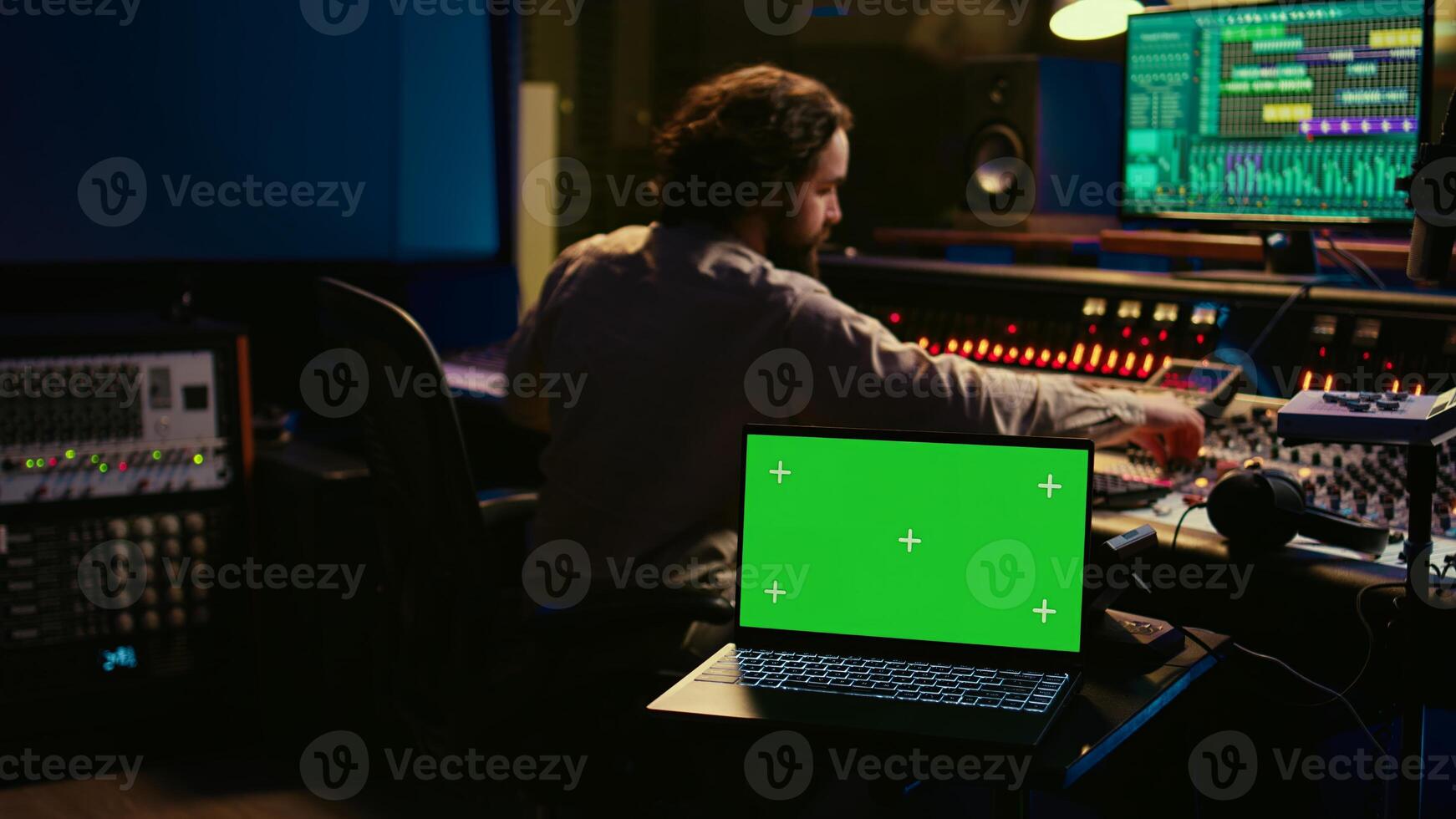 Music production engineer mixing and mastering music in control room, using laptop with greenscreen display. Skilled audio technician working with recording software on pc, pushing faders. Camera B. photo