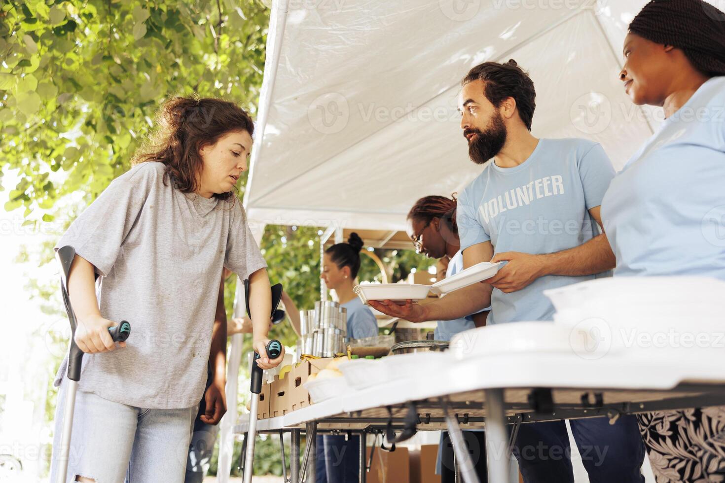 In this image, multiracial volunteers providing hunger relief support to caucasian woman on crutches. Voluntary individuals distribute free food, offer shelter, and assistance to poor and disabled. photo