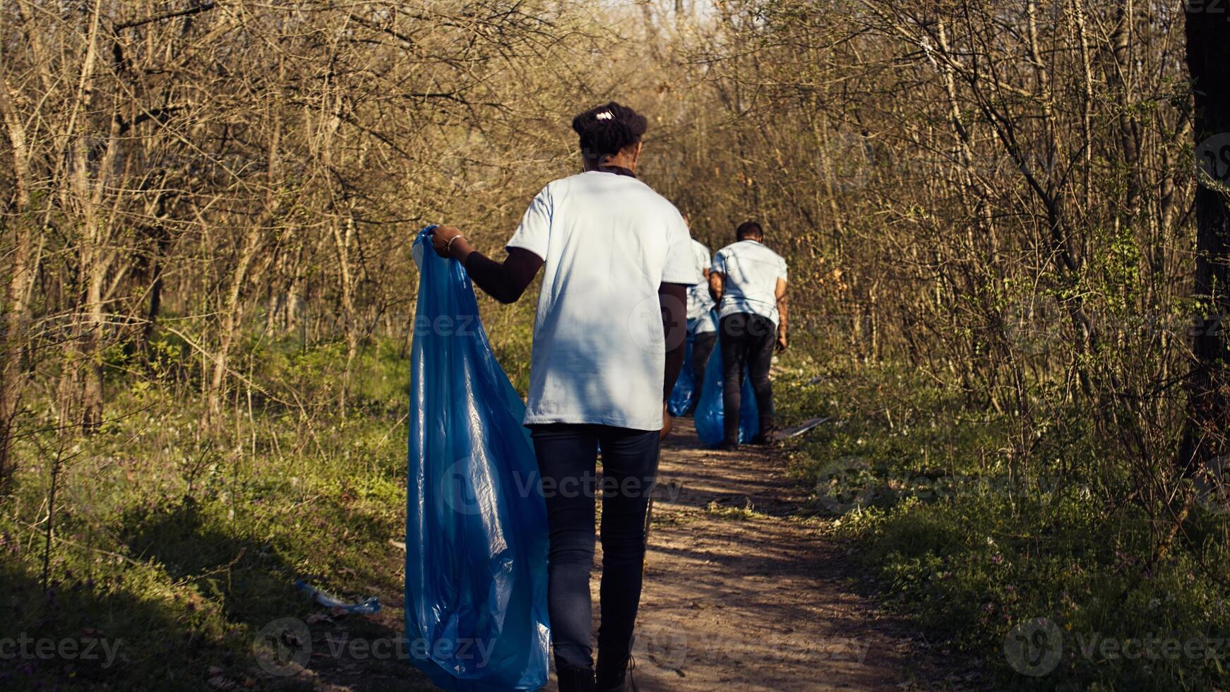 African american girl collecting rubbish in a trash bag using tongs, recycling plastic waste and picking up garbage in a forest. Young woman activist clearing the woods from junk. Camera B. photo