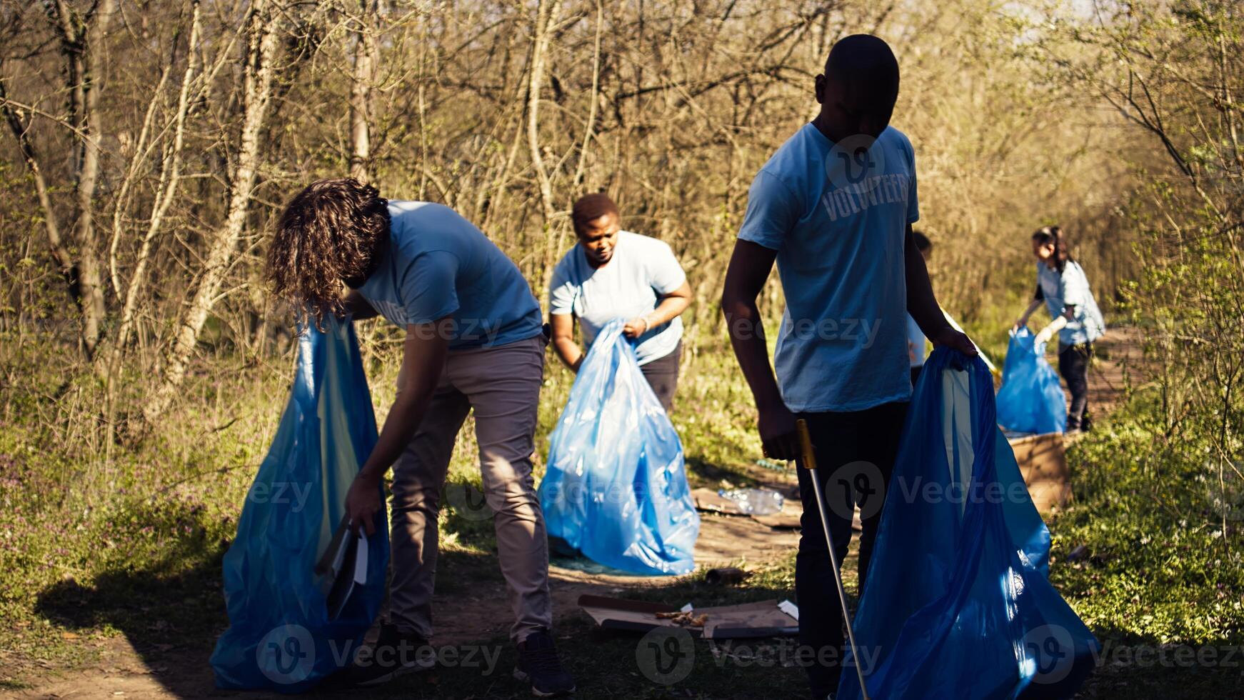 Diverse men volunteers pick up rubbish and plastic trash with tongs, working to combat illegal dumping and preserve natural forest environment. Activists volunteering for litter cleanup. Camera B. photo