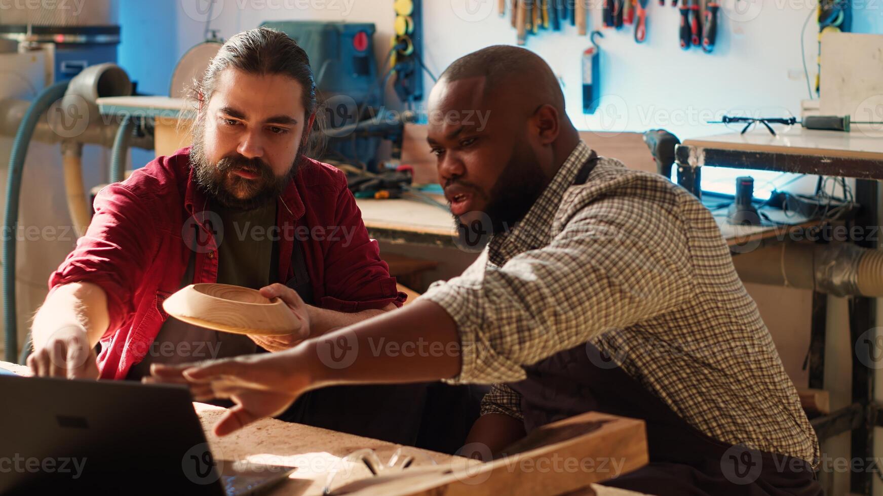Woodworker looking over blueprints on laptop, brainstorming with african american apprentice. Team of manufacturers discussing furniture assembly schematics on notebook in studio, camera A photo