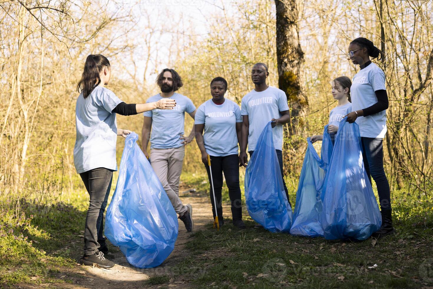 Group of nature activists collaborating to clean the woods area from trash, prepared with instruments and disposal bags. Diverse people fighting illegal dumping, ecosystem responsibility. photo