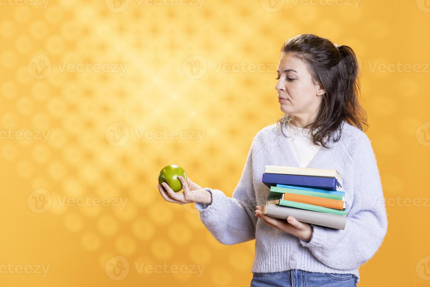 Woman with stack of books in hands enjoying fresh apple, living healthy, isolated over studio background. Bookworm holding pile of novels and green fruit, enjoying bio snack photo