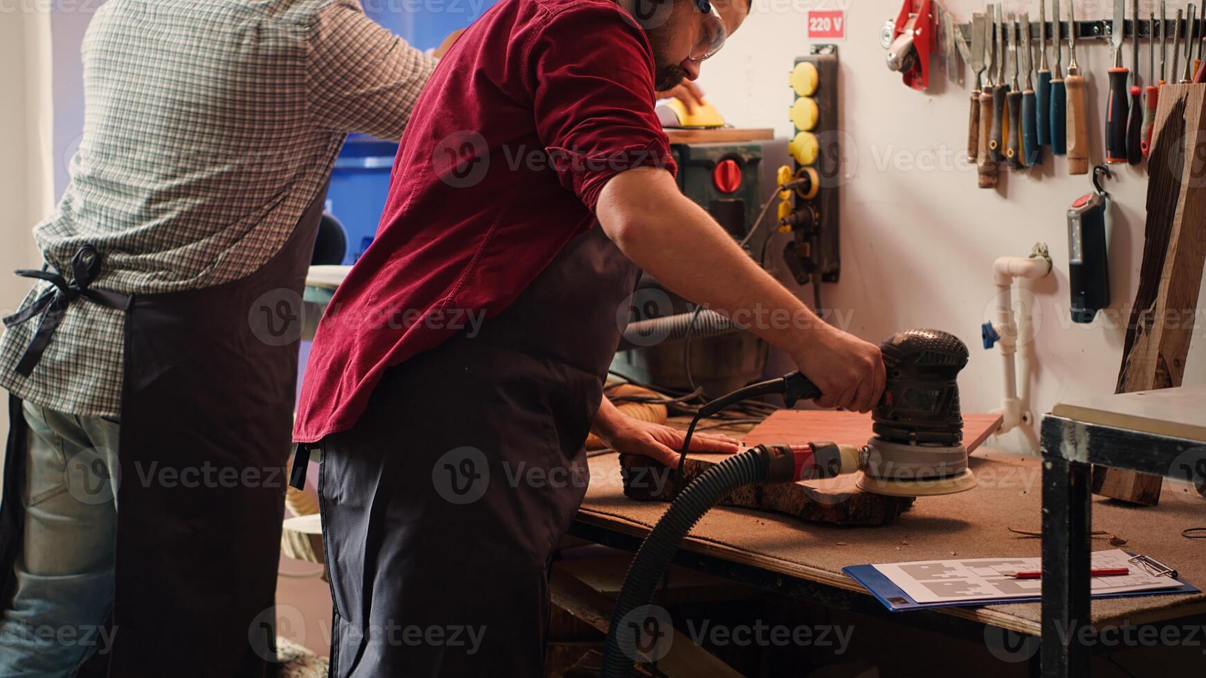 Craftsperson at work consulting blueprint and using orbital sander with sandpaper on lumber. Woodworking expert in carpentry shop uses angle grinder on wood after checking schematics, camera A photo