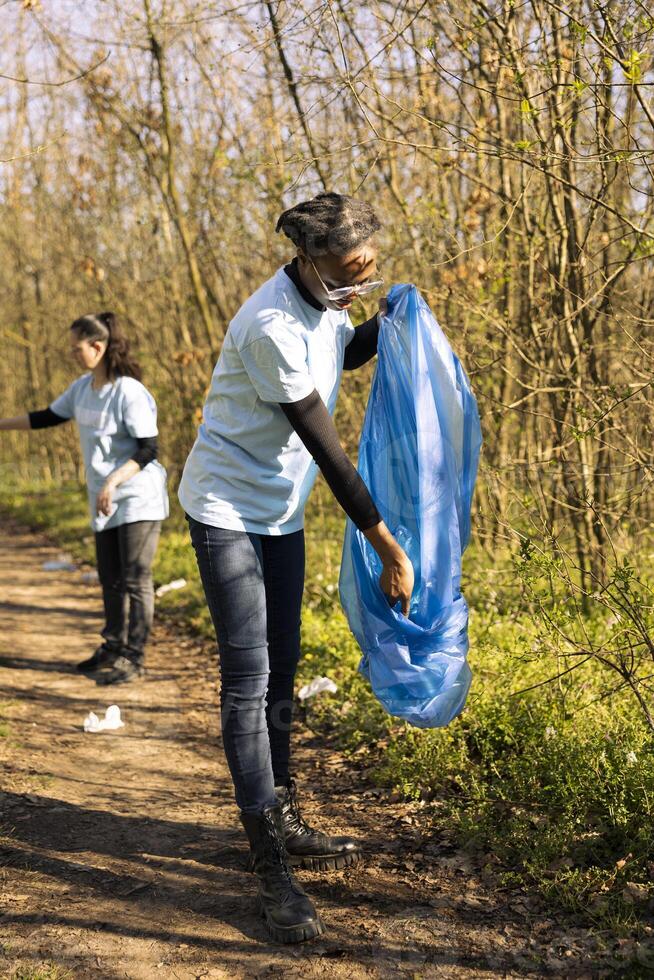 Woman volunteer grabbing junk and plastic waste with a claw tool, clearing natural ecosystem of garbage. African american girl doing voluntary work against illegal dumping. photo