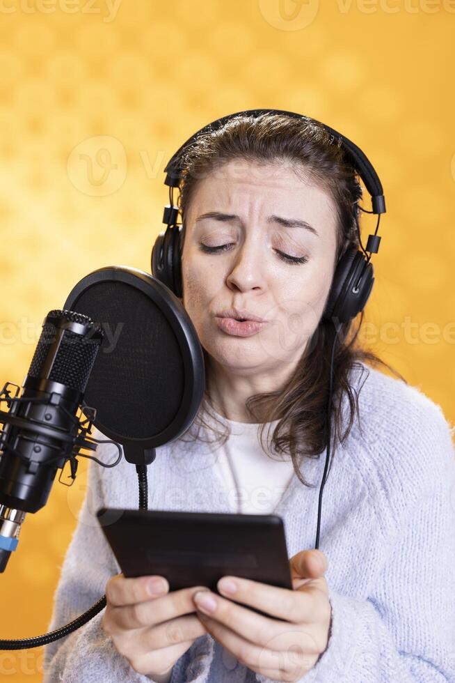 Woman frowns while doing voiceover reading of ebook on ereader to produce audiobook using dramatic acting. Narrator glowers, portraying character, recording novel using digital tablet, studio background photo