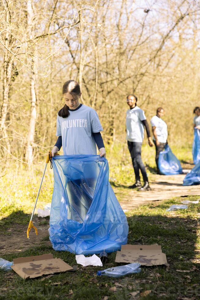 Community service volunteer reduces waste and cleans up forest setting, collecting garbage in blue trash bags. Young kid helps environment conservation by disposing of plastic waste. photo