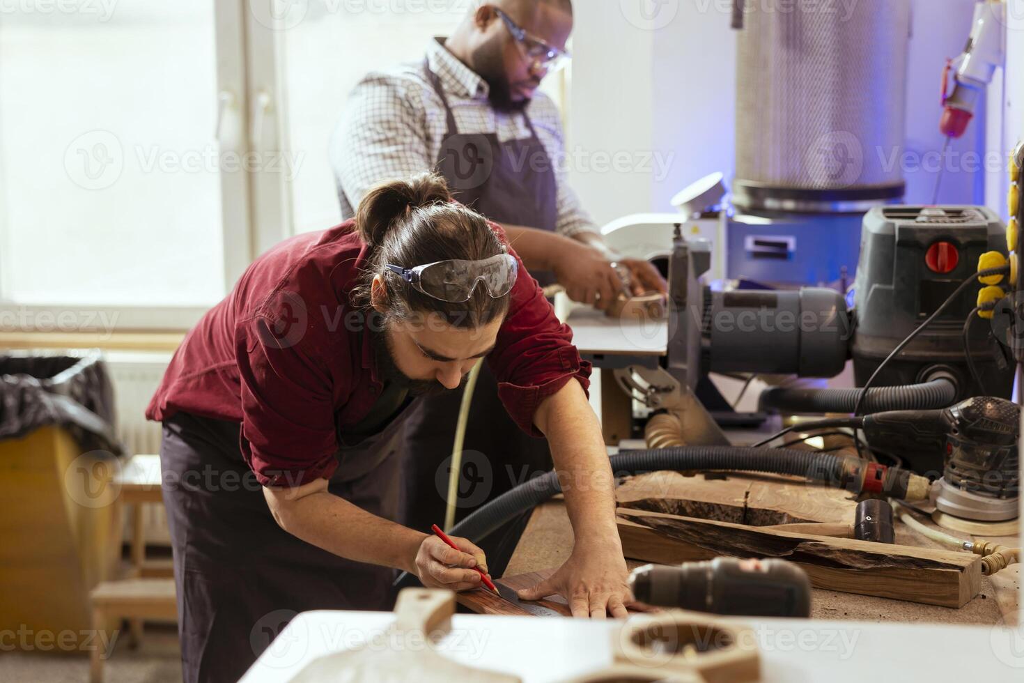 Woodworker using ruler to take measurements of wood plank with african american coworker. Creative person drawing with pencil on wooden board to determine where to make cut, helped by apprentice photo