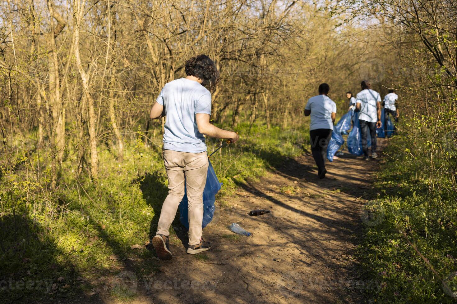 Young nature activist collecting junk with a long claw tool and garbage bag, grabbing plastic waste and trash to clean the woodland. Male volunteer does community service, global pollution. photo