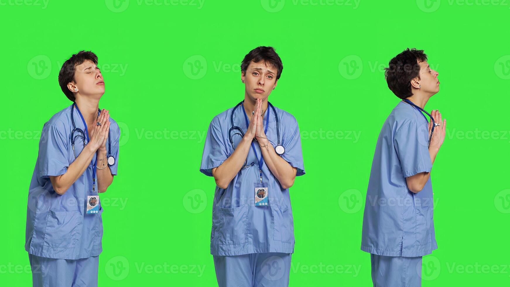 Medical assistant being hopeful praying for good luck against greenscreen, holds hands in a prayer sign and having belief in spirituality. Young christian nurse with faith in meditation. Camera B. photo