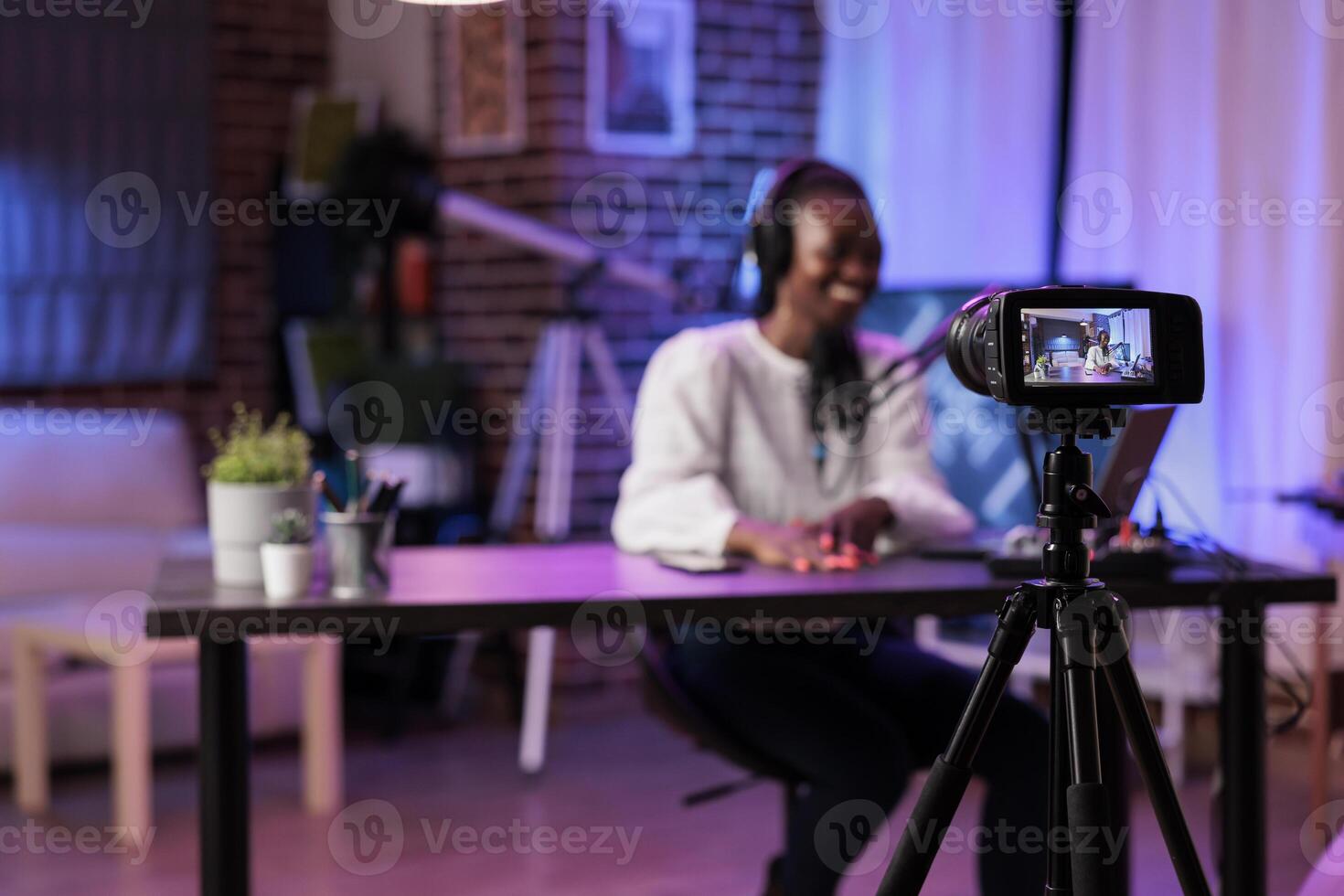 Focus shot on modern camera recording vlogger in blurry background talking about family and relationships subjects. Woman producing online show using high quality equipment in rgb lights studio photo