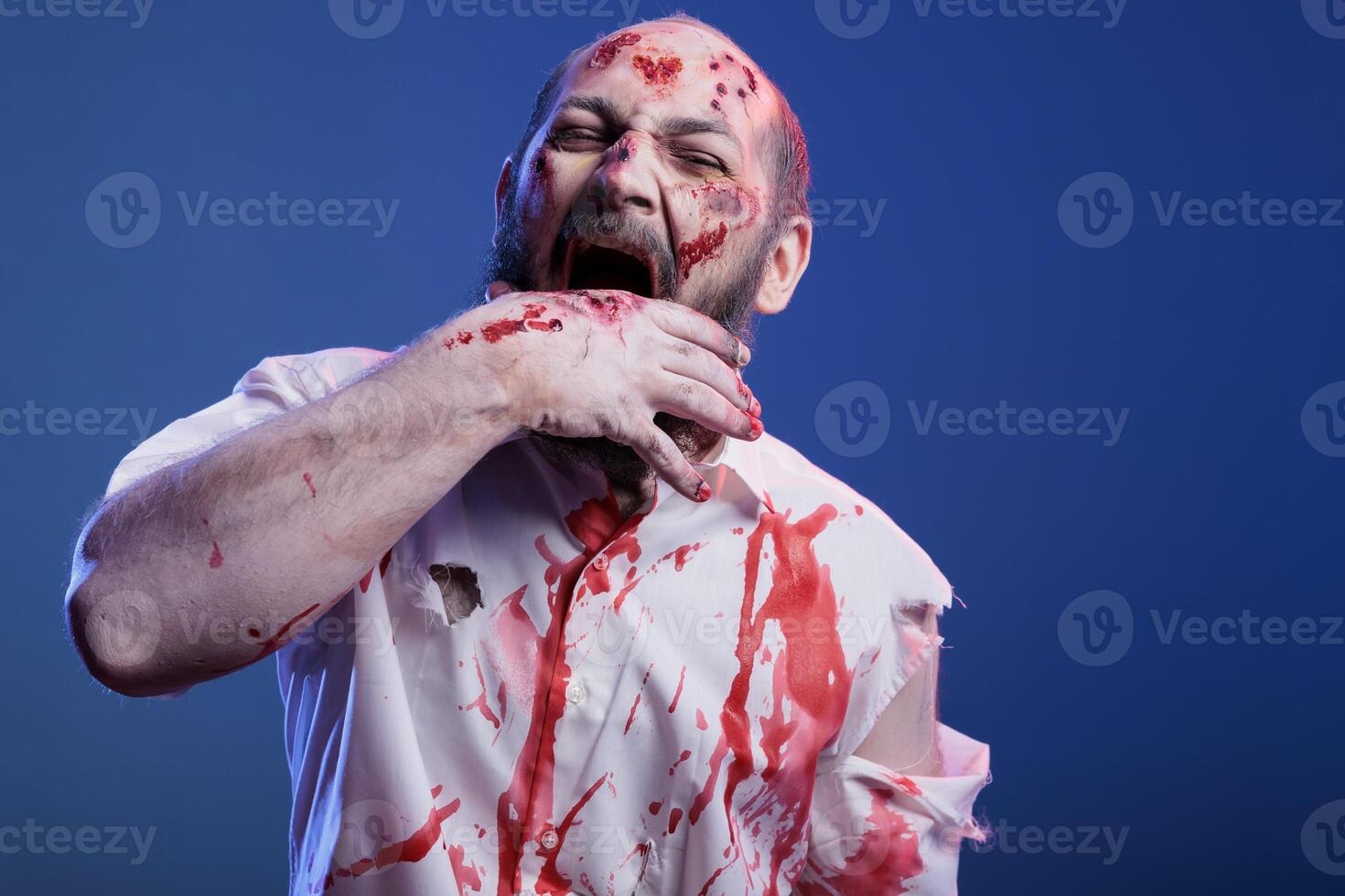 Portrait of devilish maniac zombie with blood on face and clothes biting his hand, feeling hungry for flesh. Crazy undead monster chewing on own arm, isolated over studio background photo