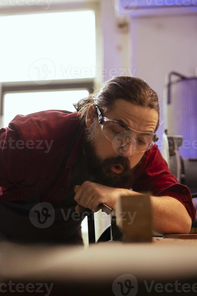 Sculptor blowing sawdust from workbench after shaping raw timber using chisel and hammer, creating wood art. Artist making handmade wood sculptures, making precise grooves in lumber block with tools photo