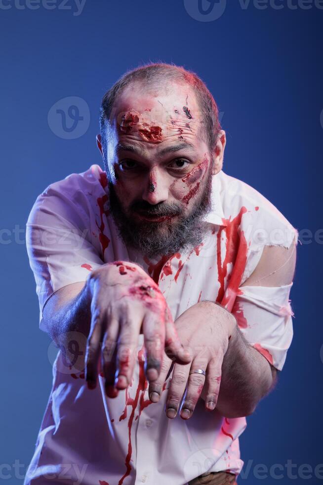 Portrait of man dressed as terrifying creepy zombie covered in blood for halloween party. Person wearing scary makeup, pretending to be undead monster with scars, studio background photo