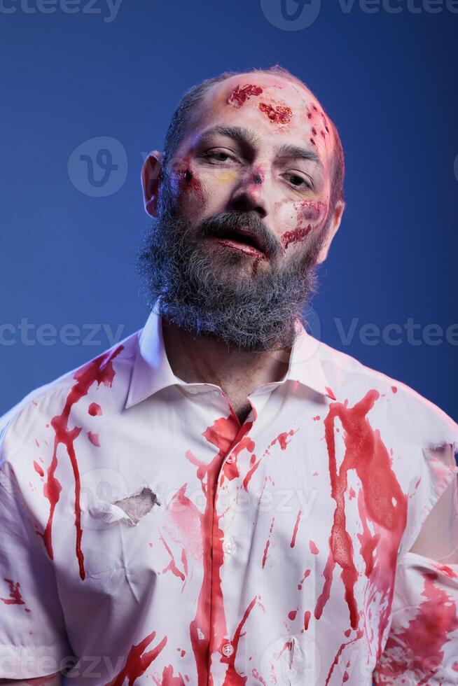 Portrait of actor portraying zombie character in horror movie with blood on face and clothes. Man dressed as undead creature for upcoming thriller film, wearing fake wounds makeup, studio background photo