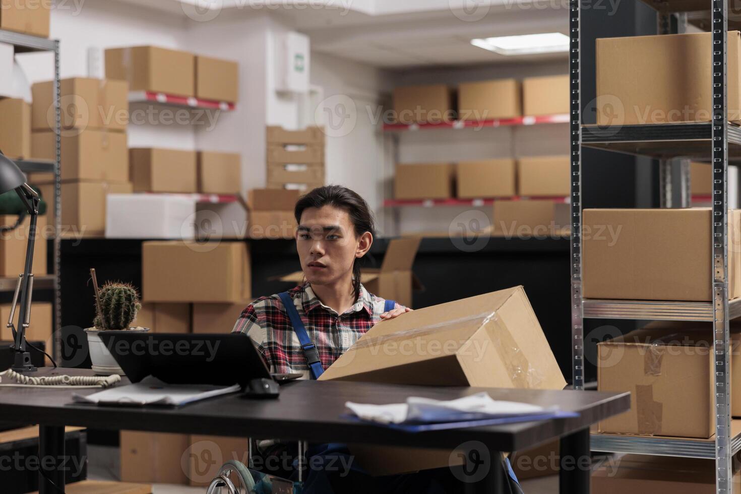 Warehouse employee in wheelchair processing order on laptop while holding cardboard box ready for shipment. Storehouse asian young man delivery manager working in disability friendly workplace photo