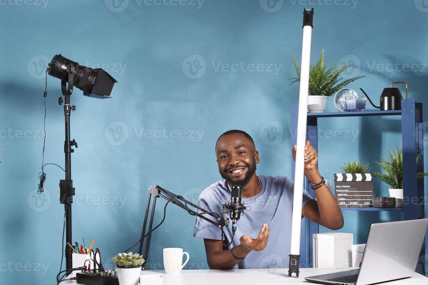 Smiling blogger holding tube light equipment and explaining features while recording for online channel. Vlogger showing led lamp for photography studio and live streaming product review photo