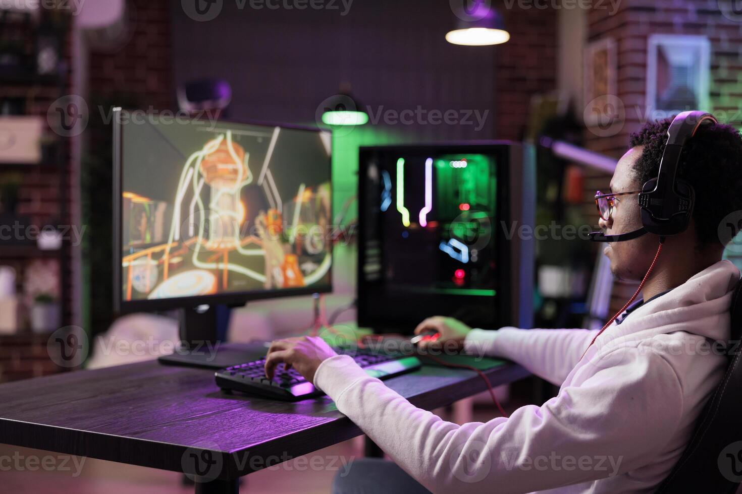 Man in brick wall living room playing games on gaming PC at computer desk, enjoying day off from work. Gamer battling enemies in online multiplayer shooter from neon lights ornate home photo