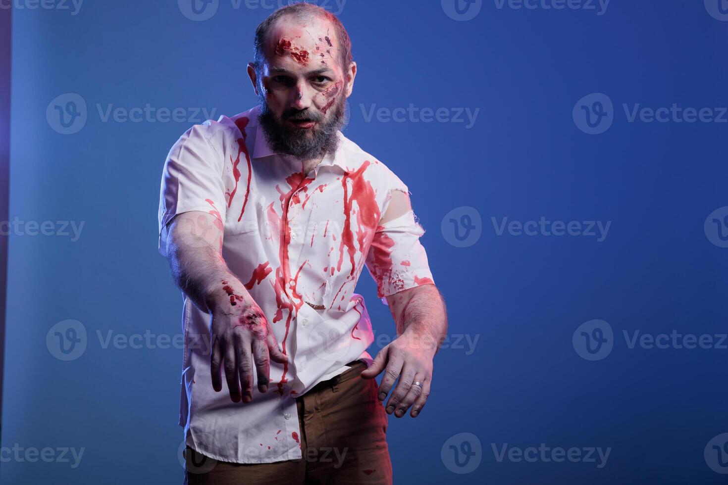Portrait of evil reanimated zombie ready to attack and eat brains, infected by virus. Terrifying undead creature covered in blood and with rotten skin limping towards camera, studio background photo