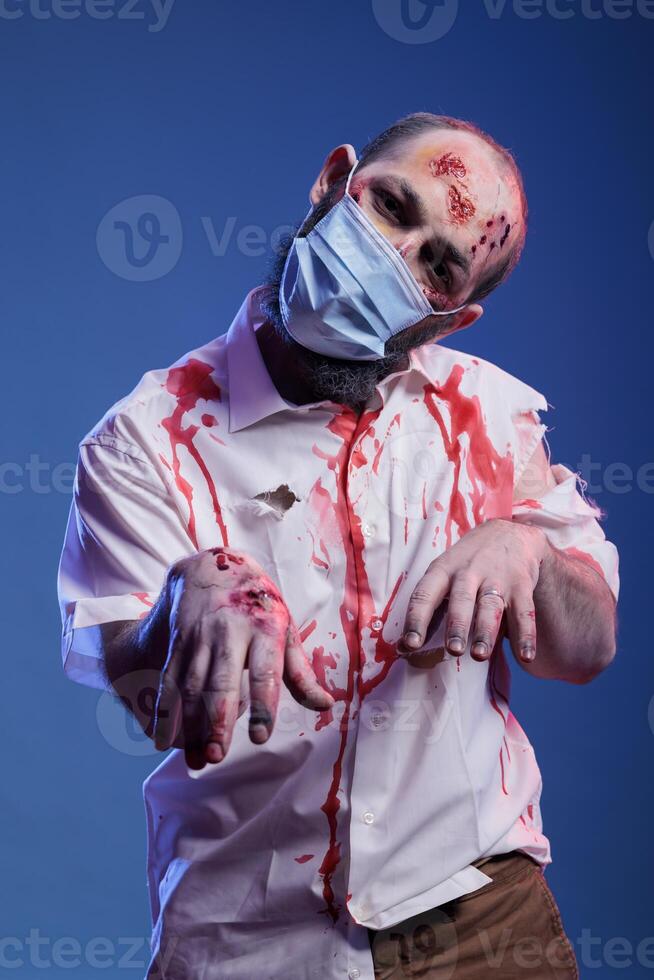 Portrait of actor playing zombie in horror movie wearing face mask during coronavirus outbreak. Man dressed as reanimated corpse for upcoming thriller film, infected with covid, studio background photo